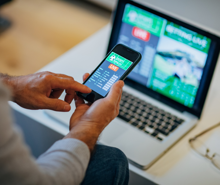 Picture of someone using their phone and laptop to place a bet on a sports game. 
