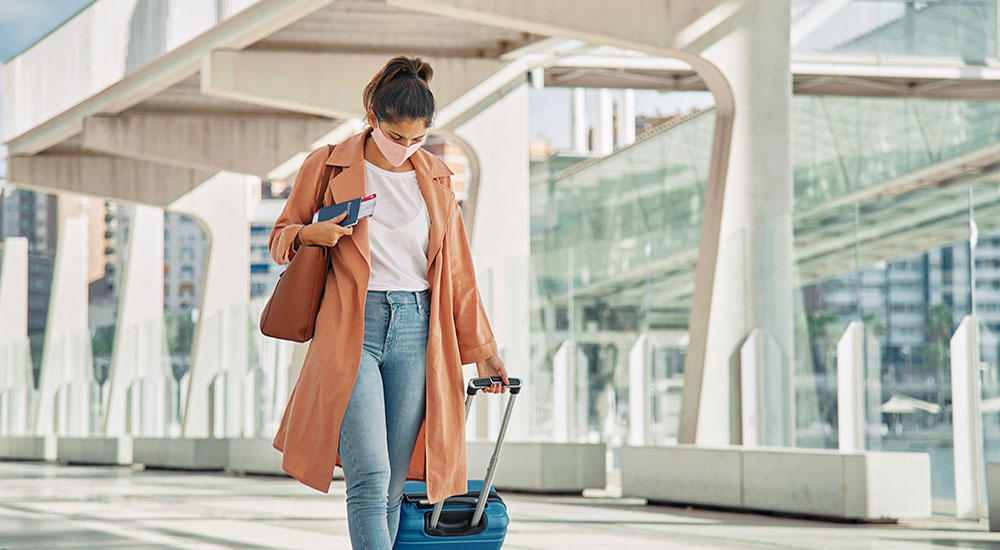 Woman wheeling a blue suitcase outside an airport. She has her head down with her brown hair tied up and a pink face mask on. She's carrying a passport with a ticket tucked out of it. 