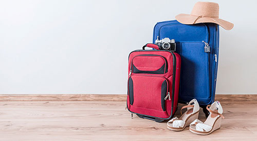 A red and blue suitcase stand together in a space with a white wall and wooden floor. A camera, sun hat and wedge heels rest on and next to the suitcases. 