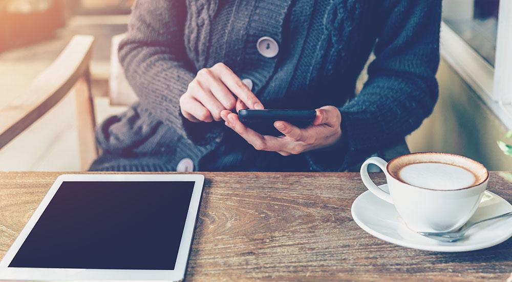Image of someone holding a smart phone at a table, they are sat down with a coffee in front of them and an electronic tablet on the table. 